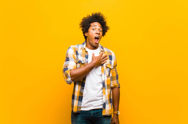 young black man feeling shocked, astonished and surprised, with hand on chest and open mouth, saying who, me? against orange wall - Фото, изображение