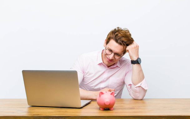 young red head businessman working in his desk with a piggy bank - Foto, afbeelding