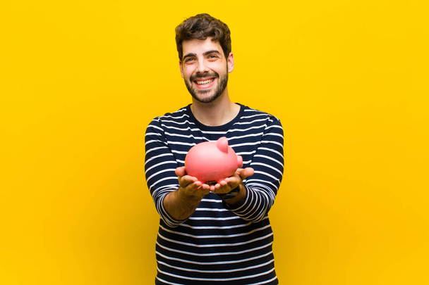 young handsome man holding a piggy bank against orange backgroun - Zdjęcie, obraz