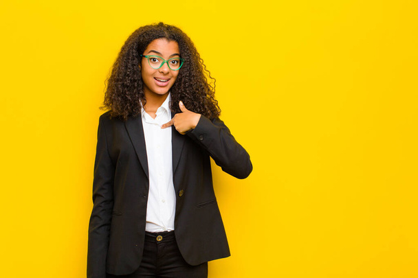 black business woman looking happy, proud and surprised, cheerfully pointing to self, feeling confident and lofty against orange wall - Foto, Imagen
