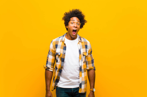 young black man feeling terrified and shocked, with mouth wide open in surprise against orange wall - Фото, изображение