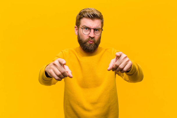 young blonde man pointing forward at camera with both fingers and angry expression, telling you to do your duty against orange wall - Fotoğraf, Görsel