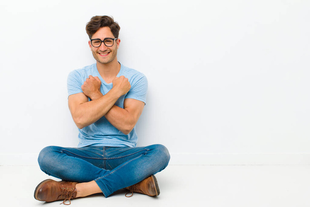 young handsome man smiling cheerfully and celebrating, with fists clenched and arms crossed, feeling happy and positive sitting on the floor - Foto, immagini