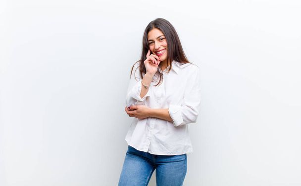 young pretty woman smiling, enjoying life, feeling happy, friendly, satisfied and carefree with hand on chin isolated against white wall - Photo, Image