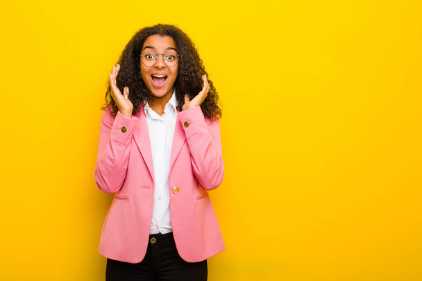 black business woman feeling shocked and excited, laughing, amazed and happy because of an unexpected surprise against orange wall - Foto, afbeelding