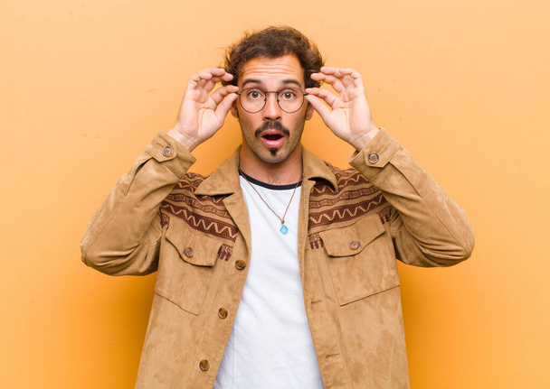 young handsome man feeling shocked, amazed and surprised, holding glasses with astonished, disbelieving look against orange wall - Fotoğraf, Görsel