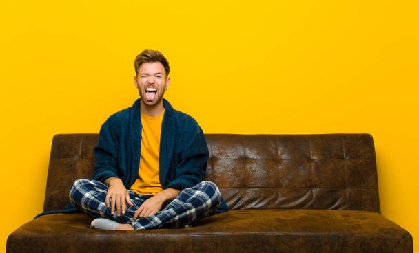 young man wearing pajamas with cheerful, carefree, rebellious attitude, joking and sticking tongue out, having fun . sitting on a sofa - Фото, зображення