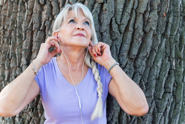 Senior lady putting headset in ears for listening music at park - Foto, Imagem