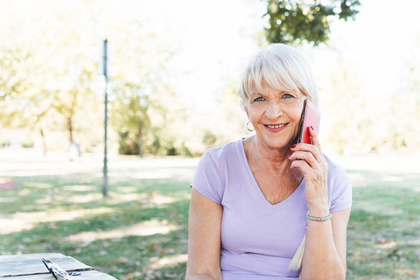 Cheerful aged woman taking rest at park after running and talking by smartphone - Фото, изображение