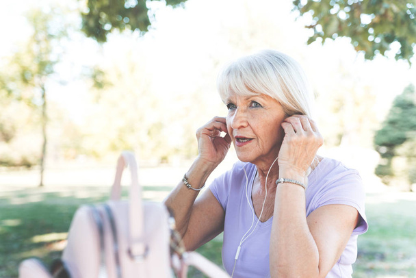 Η ηλικιωμένη κυρία βάζει ακουστικά στα αυτιά για να ακούει μουσική στο πάρκο. - Φωτογραφία, εικόνα