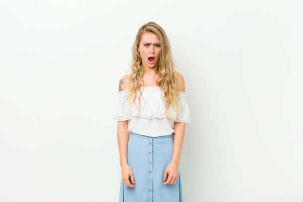 young blonde woman feeling terrified and shocked, with mouth wide open in surprise against white wall - Photo, Image