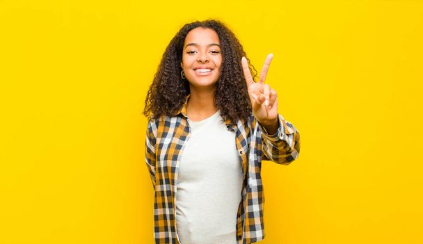 nuori kaunis afrikkalainen amerikkalainen nainen hymyilee ja näyttää onnelliselta, huoleton ja positiivinen, eleenä voitto tai rauha yhdellä kädellä vastaan keltainen seinä
 - Valokuva, kuva
