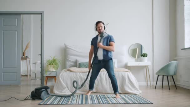 Happy young man in headphones singing in vacuum cleaner dancing cleaning home - Filmagem, Vídeo