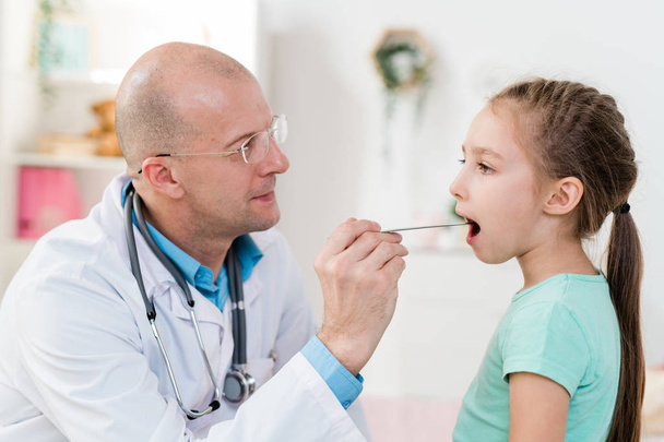 krankes kleines Mädchen hält Mund offen, während Arzt im weißen Kittel ihre Halsschmerzen mit medizinischem Werkzeug untersucht - Foto, Bild