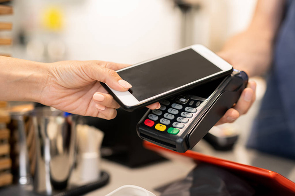 Young woman using her mobile phone for contactless payment while holding it over electronic machine held by barista - Photo, Image