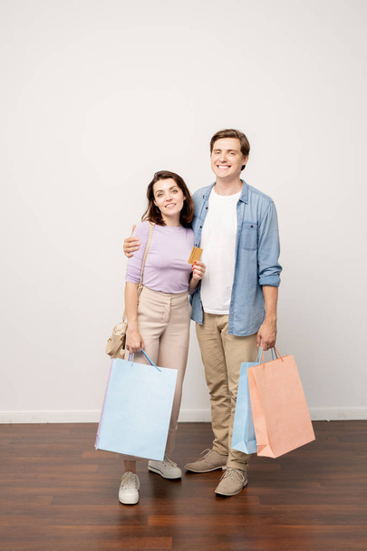 Happy couple of young shoppers with paperbags and credit card standing by wall in isolation in studio - Photo, Image