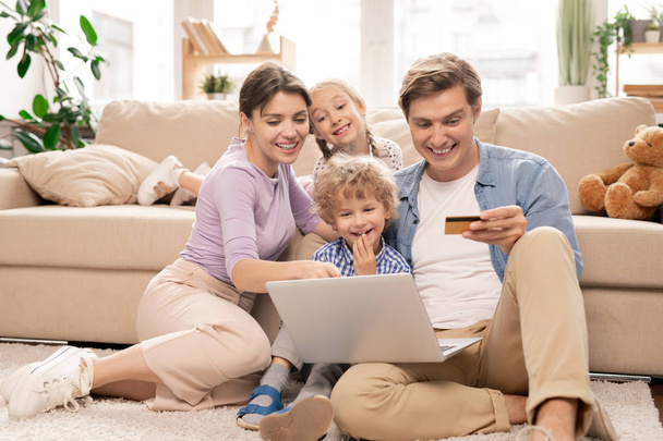 fröhliche junge vierköpfige Familie sitzt auf dem Boden des Wohnzimmers und surft im Online-Shop, während sie sich etwas aussucht - Foto, Bild
