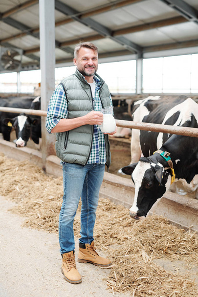 Zdrowy młody rolnik z dzbankiem mleka patrząc na ciebie stojąc w gospodarstwie mleczarskim przez oborę - Zdjęcie, obraz