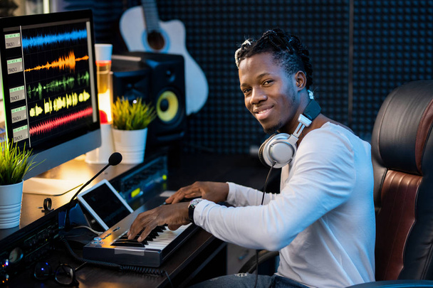 Boldog fiatal zenész fülhallgatóval a nyakán néz rád, miközben dolgozik az új zene munkahelyi stúdióban - Fotó, kép