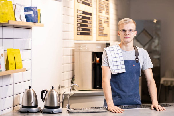 Young male barista in uniform looking at you while standing by table with two electric teapots and coffee machine on background - Fotografie, Obrázek
