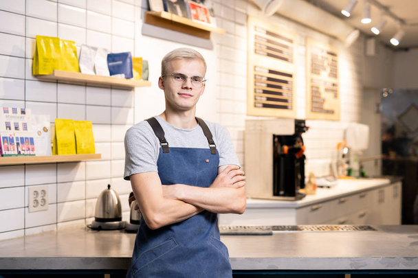 Jeune barista à bras croisés ou propriétaire d'une cafétéria debout à table sur fond d'équipement de travail et de paquets de café
 - Photo, image