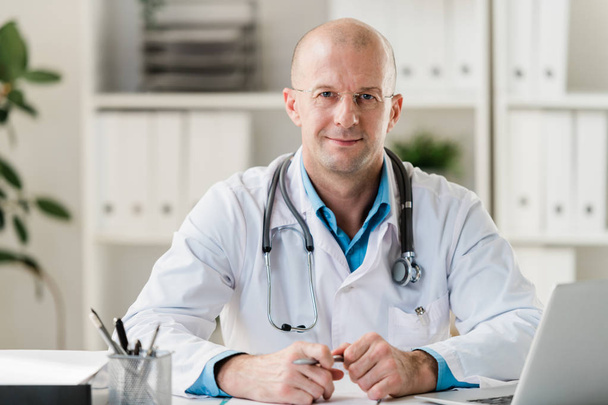 Médecin professionnel chauve avec stylo assis par bureau dans un cabinet médical ou un hôpital et effectuant son travail
 - Photo, image