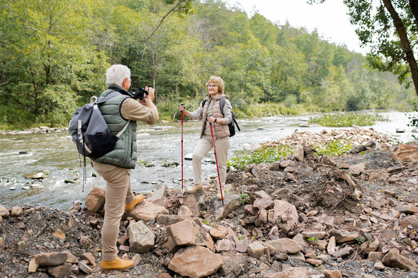 Homme mature actif avec sac à dos et appareil photo photographiant sa femme heureuse avec des bâtons de trekking sur la rive de la rivière
 - Photo, image