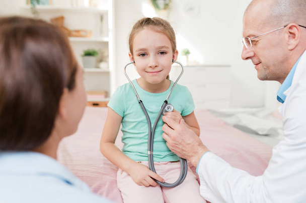 Šťastná holčička se stetoskopem poslouchá tlukot svého srdce, zatímco sedí mezi lékařem a matkou v nemocnici - Fotografie, Obrázek