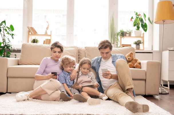 Moderne familie van jong stel en twee kinderen zitten op de vloer per bank en met behulp van hun mobiele gadgets in de woonkamer - Foto, afbeelding