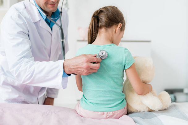 Joven pediatra en Whitecoat examinando la espalda de una niña con estetoscopio en el consultorio médico
 - Foto, imagen