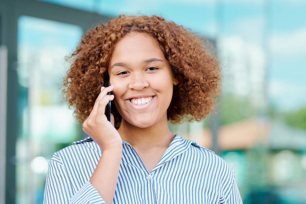 Молода весела міжкультурна бізнес-леді з кучеряво-коричневим волоссям розмовляє на смартфоні після роботи в міському середовищі
 - Фото, зображення