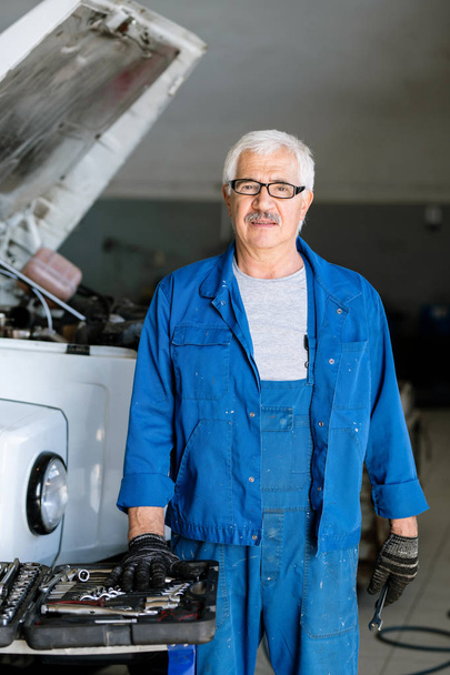 Elderly and experienced master of technical service looking at camera while standing by broken lorry in workshop - 写真・画像