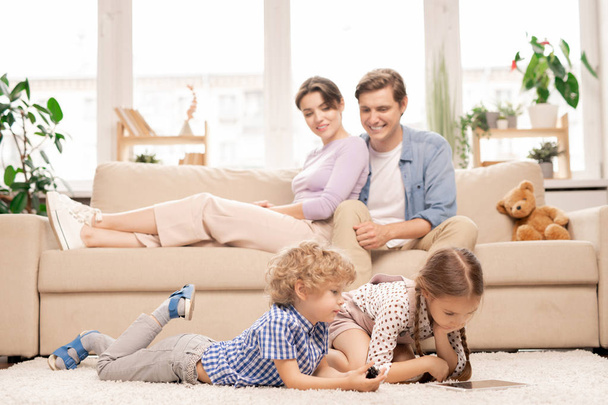Dwa słodkie dzieci w wieku podstawówki z touchpad leżące na podłodze salonu podczas oglądania kreskówek w domu - Zdjęcie, obraz