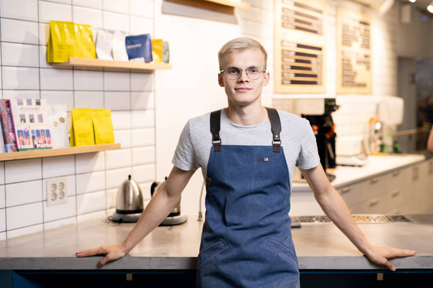 Young blond man in eyeglasses, apron and casualwear looking at you by his workplace inside cafe or restaurant - Photo, image