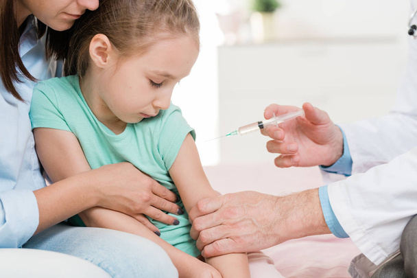 Милая маленькая девочка боится уколов, глядя на шприц в руке врача в клиниках
 - Фото, изображение