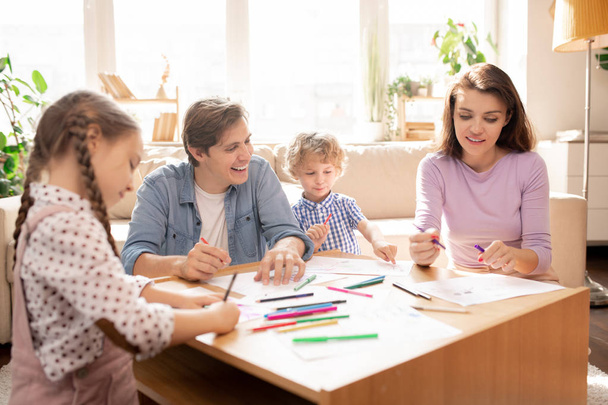 Young man laughing while looking at what his wife drew on paper with their two little kids drawing near by - Photo, Image