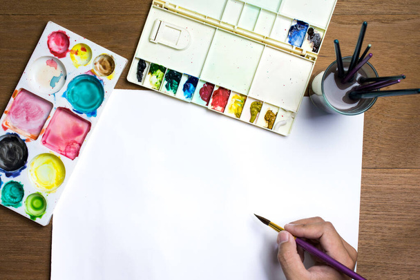 Man hand houden verfborstel op wit papier met kleurrijke aquarel en apparatuur op houten achtergrond. - Foto, afbeelding