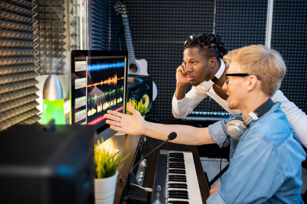 Jeune musicien africain regardant des formes d'ondes sonores sur écran d'ordinateur tandis que son collègue pointant vers l'un d'eux en studio
 - Photo, image
