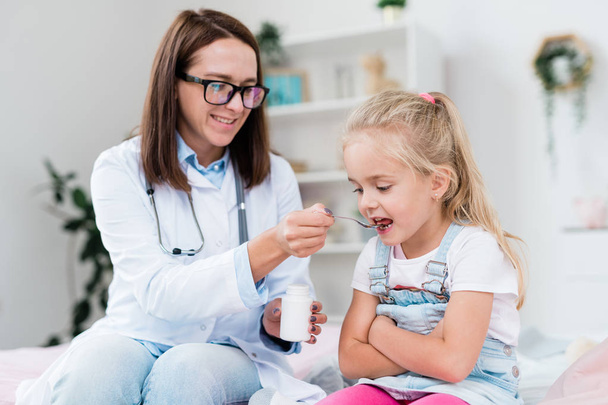 Больная маленькая девочка, принимающая лекарства из ложки, которую держит молодая женщина-врач в белом халате во время посещения больницы
 - Фото, изображение
