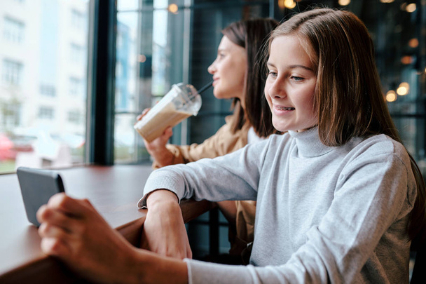 glimlachend meisje met smartphone zitten door tafel in cafe en kijken naar video met haar moeder met cocktail op de achtergrond - Foto, afbeelding