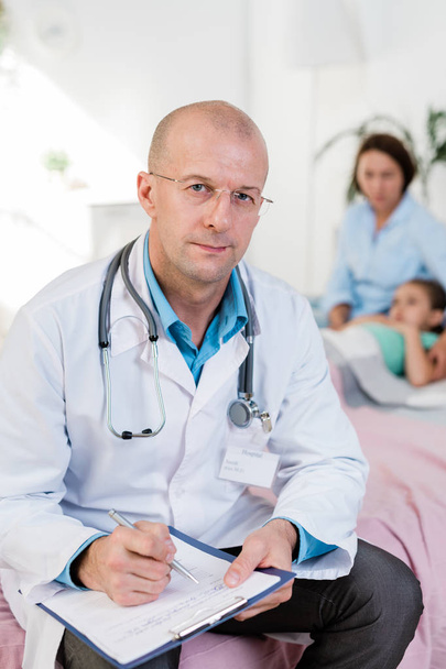 Лысый врач в белом халате с бумажными рецептами для одного из пациентов на фоне женщины и девочки
 - Фото, изображение