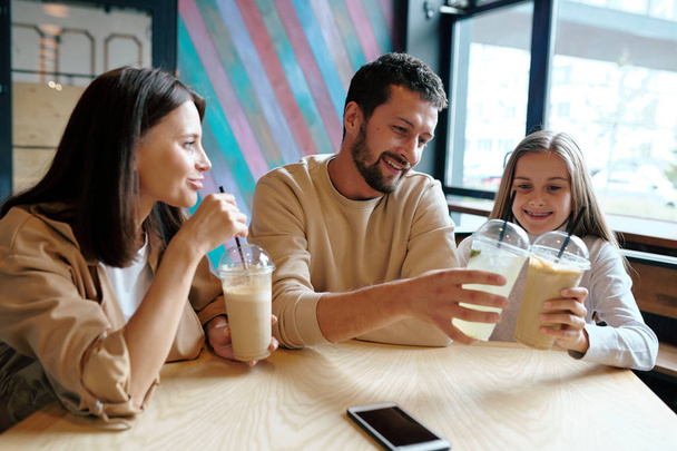 Giovane uomo e sua figlia clinking con bicchieri di plastica di cocktail di latte mentre seduto al tavolo con bella donna vicino nel caffè
 - Foto, immagini