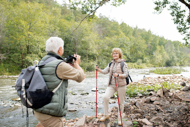 トレッキングスティックを水で石の上に立っているとアクティブな女性ハイカーは旅行中にカメラで彼女の夫を見て - 写真・画像