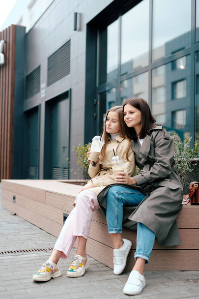 Duas fêmeas repousantes em smart casual tendo bebidas enquanto sentado ao lado do edifício moderno e desfrutando de lazer
 - Foto, Imagem