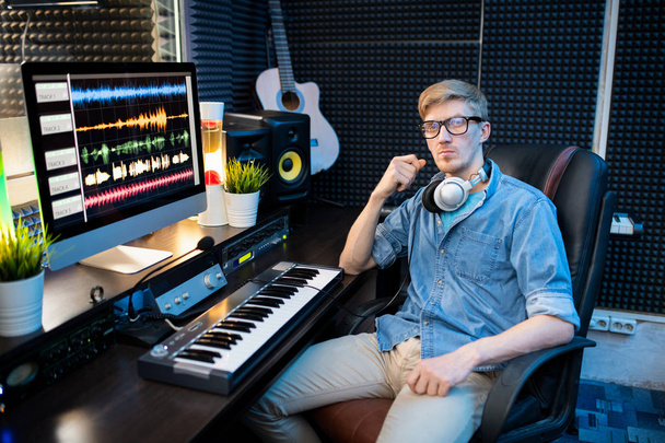 Komoly fiatalember alkalmi ruhák ül karosszékben íróasztal mellett számítógép monitor és billentyűzet, miközben keverés hangok stúdióban - Fotó, kép