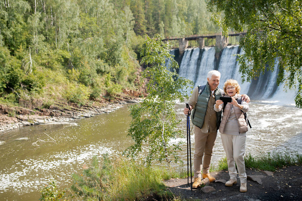 Веселая пожилая пара в активной одежде делает селфи на фоне водопадов, стоя на берегу реки в лесу
 - Фото, изображение