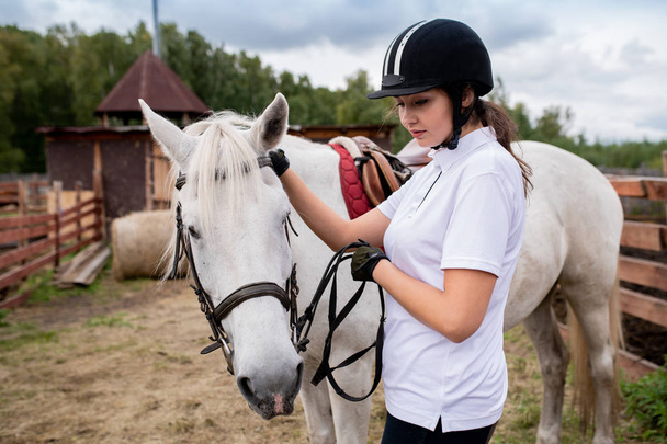 Menina ativa em capacete equestre e camisa de pólo branco e seu cavalo de corrida movendo-se para baixo campo em ambiente rural
 - Foto, Imagem