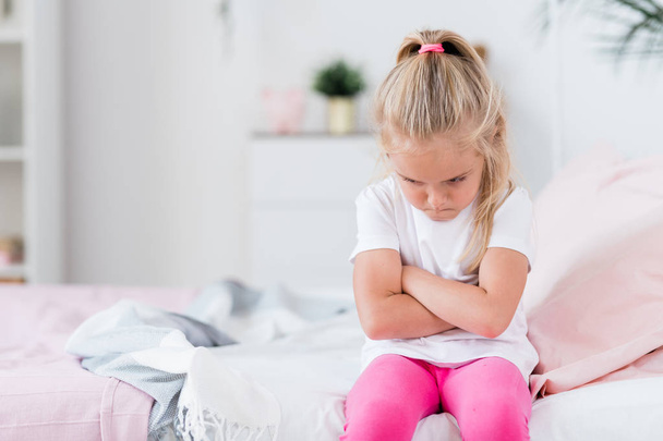 Loukkaantunut tai kyllästynyt lapsi satunnaisissa vaatteissa ristikkäin rinnassa istuessaan sohvalla kotona
 - Valokuva, kuva