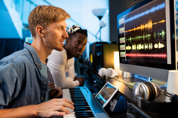 junger Mann mit Klavierbrett sitzt vor dem Computerbildschirm und betrachtet Klangwellenformen mit einem Kollegen im Hintergrund - Foto, Bild