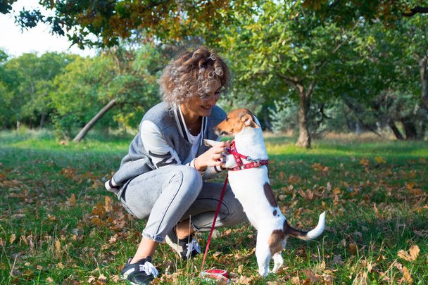 mooi blond krullend meisje speelt met haar huisdier, jack russell hond in de groene herfst park - Foto, afbeelding
